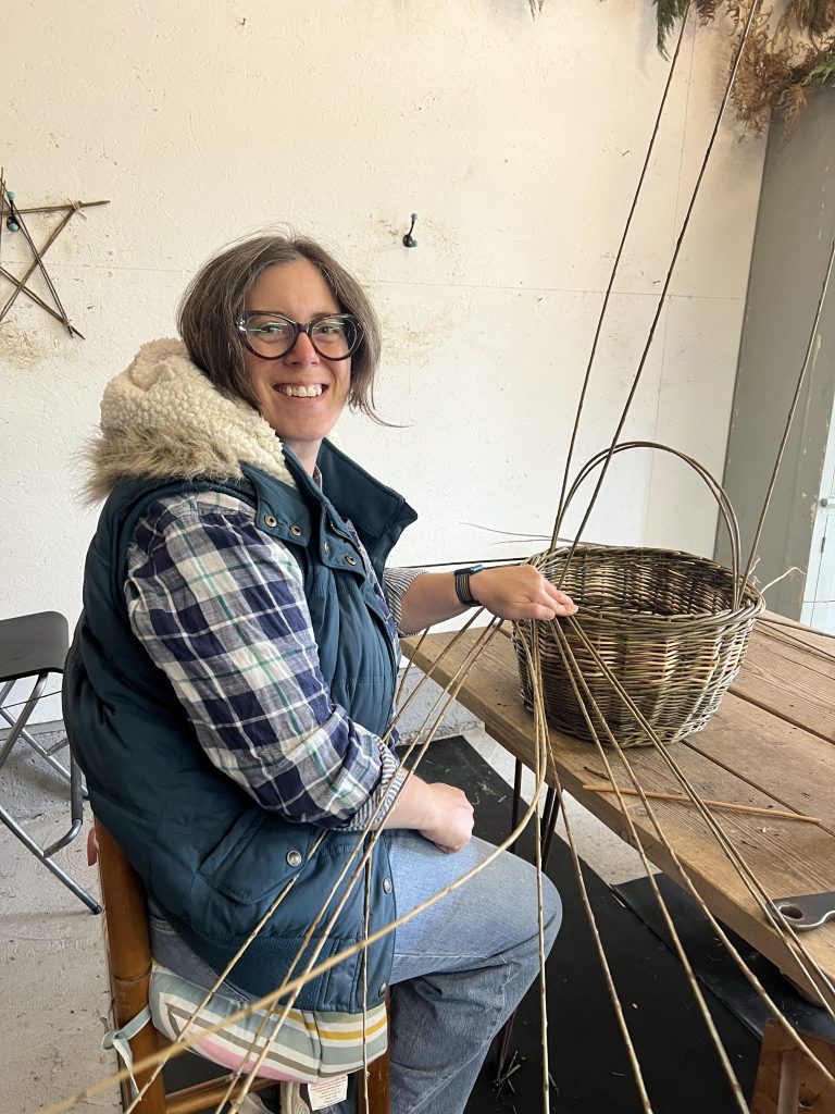 Woman in checked shirt and body warmer, sits on a stool, smiling at the camera with her hand on a basket.