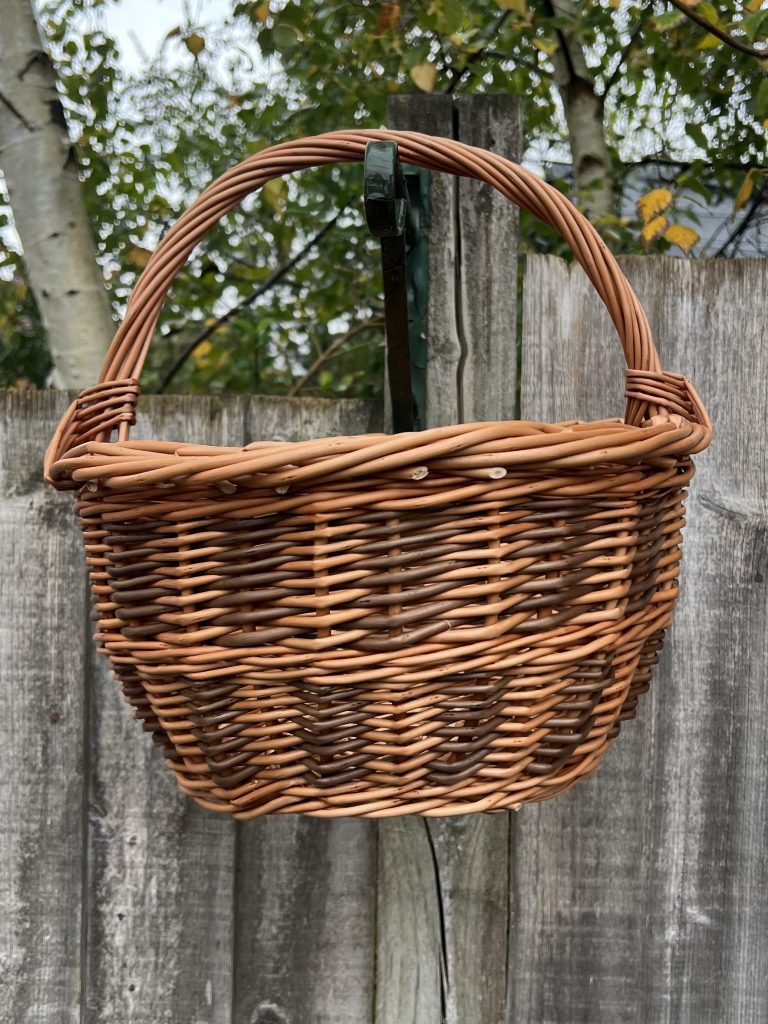 Willow basket with a chequerboard pattern hanging in front of a fence and some branches.
