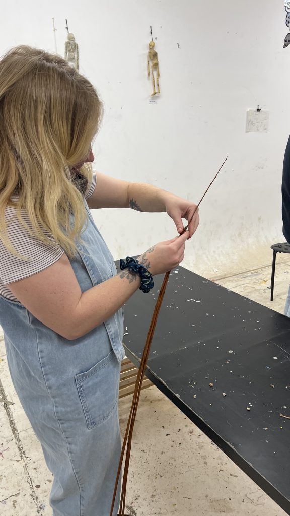 A woman with blond hair, wearing blue dungarees and a t-shirt, is standing up, facing away from the camera, holding a willow withy, in her hand.